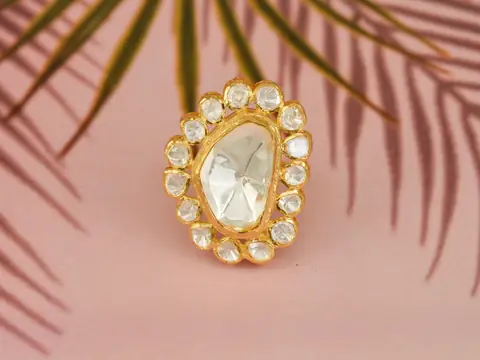 A gold ring set with a large uncut diamond on a pink ground under palm-leaf shadows