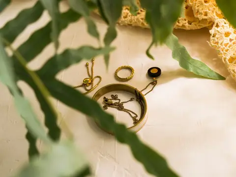 Gold bracelets, rings and fine chains laid on a pale textured surface beside a fern
